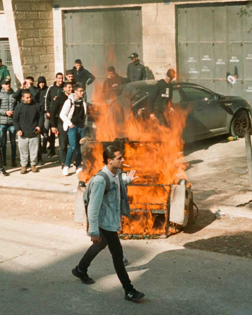 A man smoking a cigarette casually walks past a fire in Palestine, warming his hands as he goes