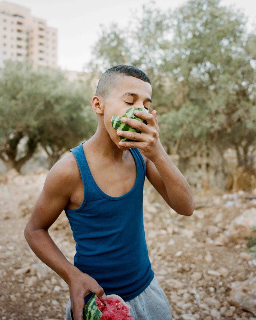 A Palestinian boy wearing a blue vest eats a melon he just picked from his farm in Beit Jala, Palestine