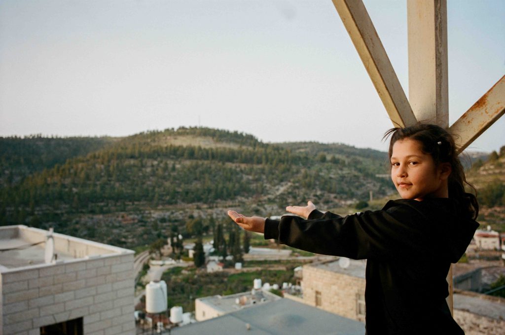 A girl stretches out her palms to check for rainfall, set against a backdrop of tree-lined hillsides in Palestine