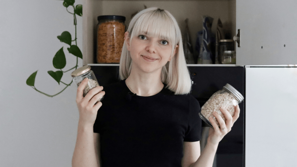 A photo of a woman with platinum blonde hair to the shoulders and bangs, holding jars in front of an organised cupboard and smiling to camera.